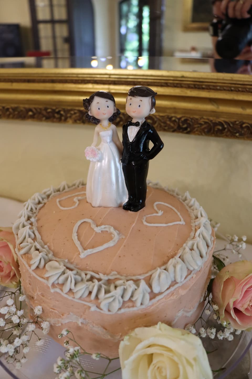 A bride and groom cake topper on a pink heart-decorated cake with roses